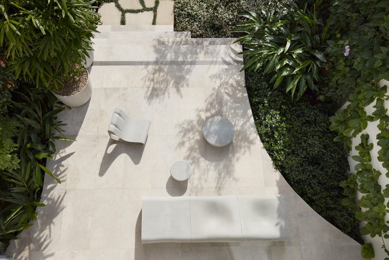 The crazy paving leads to the grey stone patio, furnished by contemporary white sofas and tables in contrast to the dark green vegetation around it