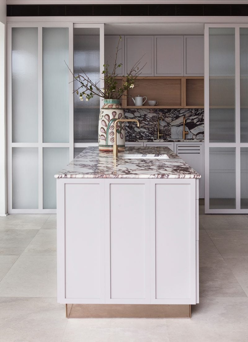 The kitchen island has white timber panels on all sides and topped by a thin calacatta viola slab. The reflective brass base gives the illusion of lightness