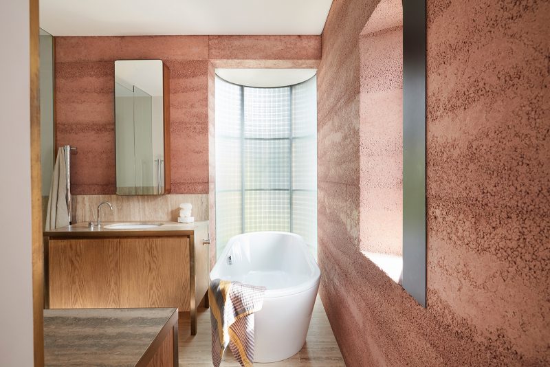 Lights dappling through mesh and translucent glass illuminates the bathtub in front of it, and highlights the textures and tones of rammed earth walls