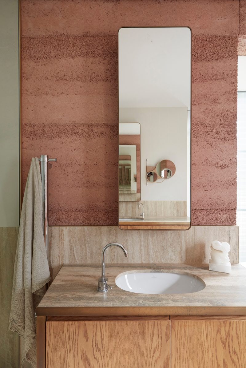 The earthy timber vanity with a similar stone splashback. The mirror above the vanity is fixed onto the pinkish earth wall behind the splashback