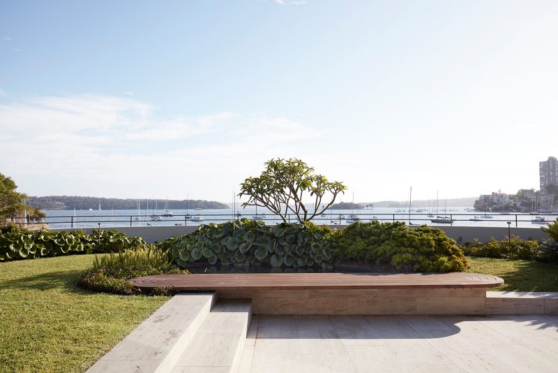Luigi Rosselli curved timber seat set into a manicured green lawn and framed by bushes, overlooking the Sydney Harbor