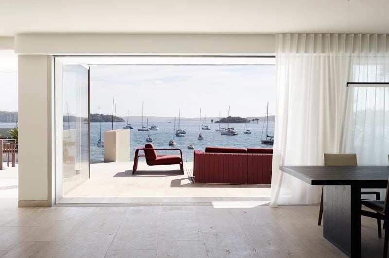 Luigi Rosselli, interior timber flooring transitions into the travertine terrace, outdoor sofa set in red overlooking sailboats in the blue Sydney harbor on a lovely warm day