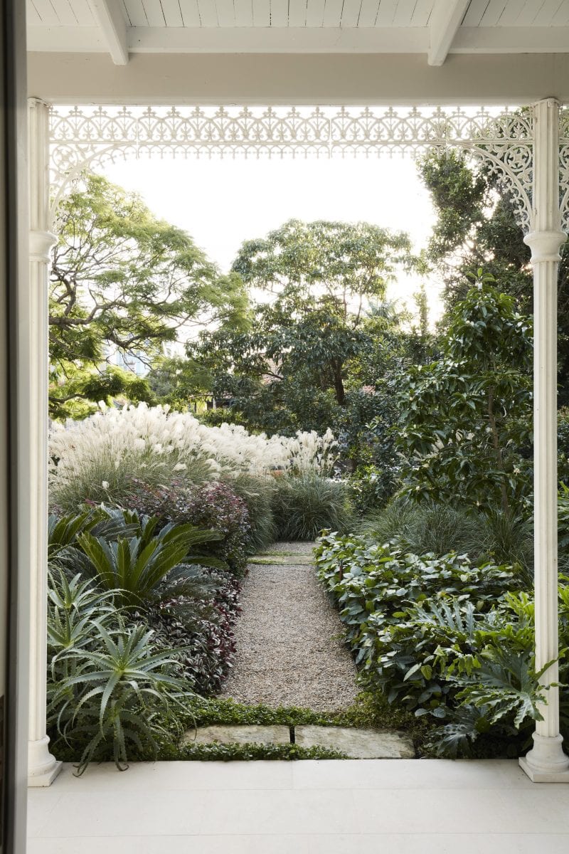 A masterfully landscaped path lined with native shrubs and tall trees, and framed by the Victorian filigree porch one stands under
